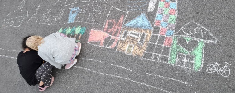 Två barn ritar hus i krita på asfalt.